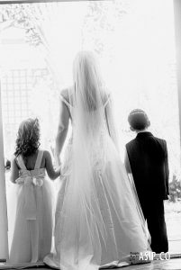 Black and white rear view of bride with page boy and flower girl holding hands captured with a cinematic, analog-inspired aesthetic by Arnold Szmerling. This manifestation emphasizes the [texture/tonal depth] and visual restraint of the Visual Life Collection.