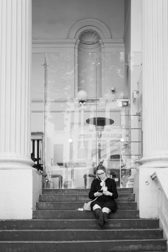A young female student checking her phone on the steps in Melbourne, a work of passion by Arnold Szmerling captured in Black and White during his Leica Q3 street photography experience.